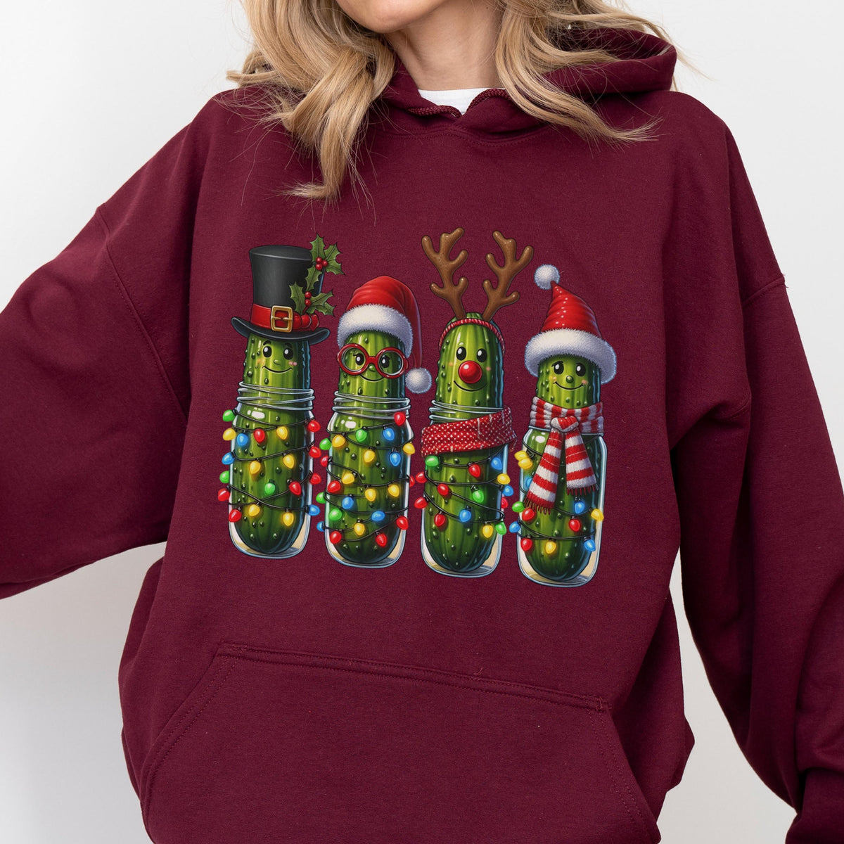 May include: A maroon hooded sweatshirt featuring a graphic print of four green pickles decorated for the holidays. The pickles wear festive accessories including a top hat, Santa hat, reindeer antlers, and a red and white striped scarf and hat.