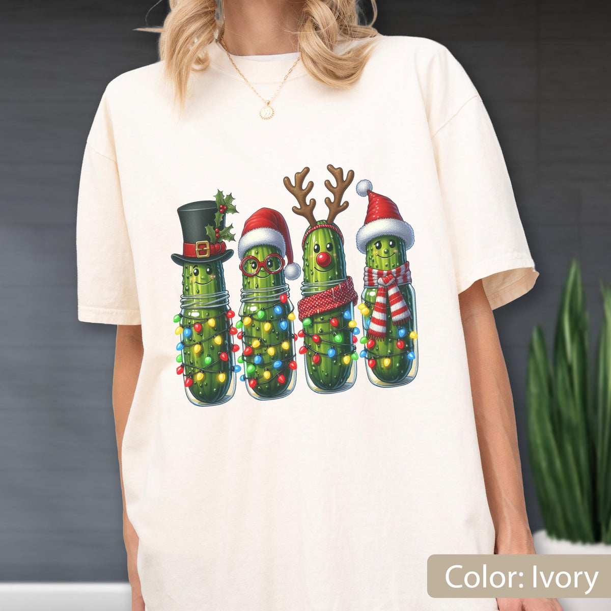 May include: An ivory t-shirt featuring a graphic print of four green pickles in glass jars. Each pickle is decorated for the holidays with items including a black top hat, a red Santa hat, reindeer antlers, and a red and white striped scarf.