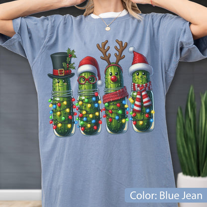 May include: A blue jean colored t-shirt featuring a graphic of four green pickles in glass jars. Each pickle is decorated for the holidays with items like a top hat, Santa hat, reindeer antlers, a red nose, scarves, and colorful string lights.