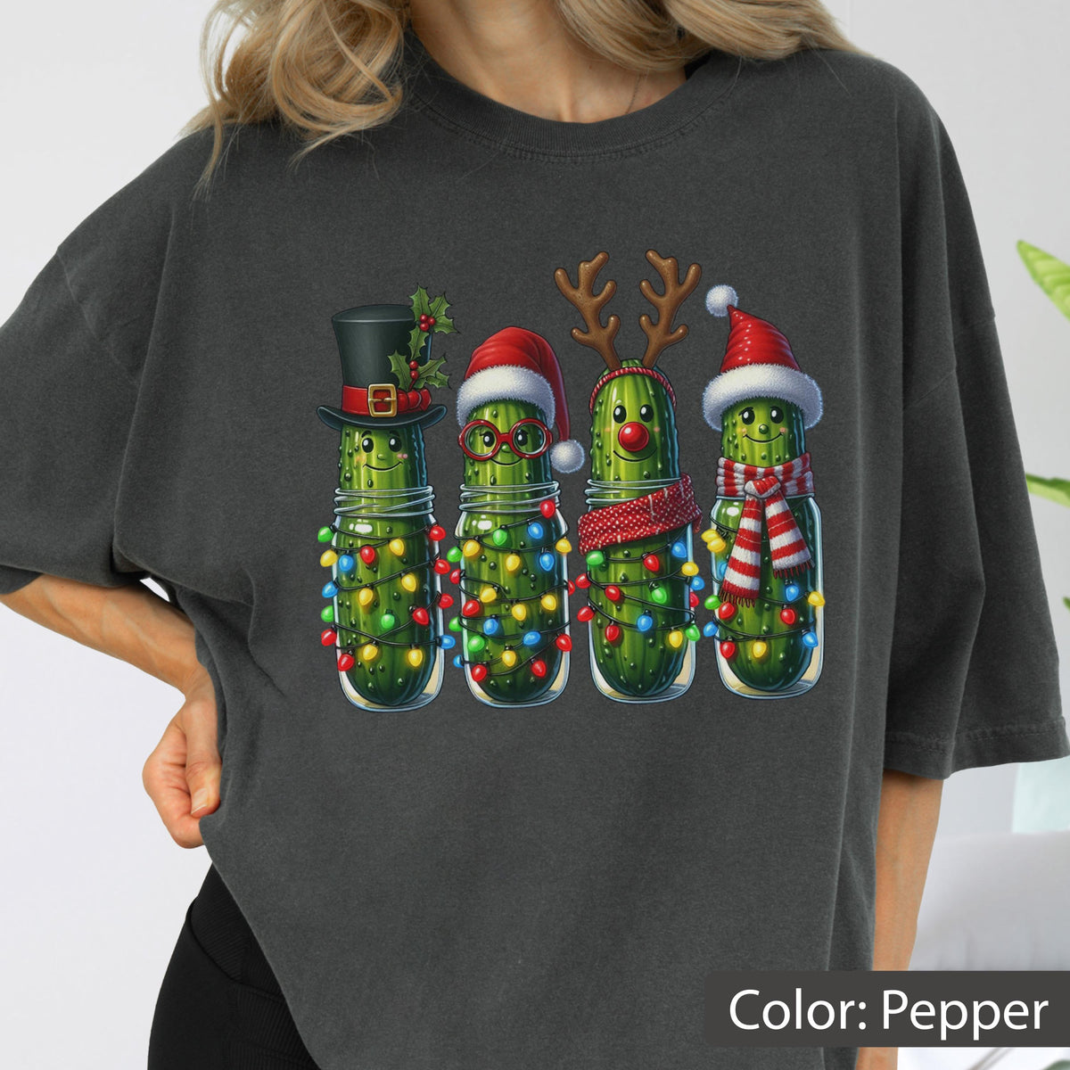 May include: A charcoal gray t-shirt featuring a graphic of four anthropomorphic pickles decorated for the holidays. The pickles are wrapped in colorful string lights and wear festive accessories including a top hat, Santa hat, reindeer antlers, and a scarf.