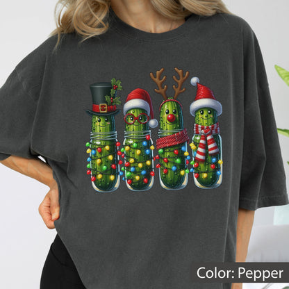 May include: A charcoal gray t-shirt featuring a graphic of four anthropomorphic pickles decorated for the holidays. The pickles are wrapped in colorful string lights and wear festive accessories including a top hat, Santa hat, reindeer antlers, and a scarf.