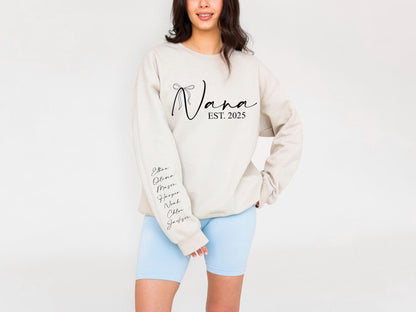 May include: Beige sweatshirt with the word "Nana" in a script font, with "EST. 2025" below. The sleeve has names written in a script font. The person is wearing light blue shorts.
