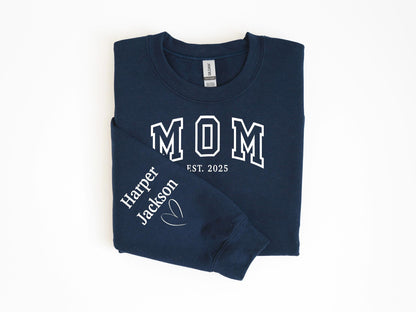 May include: Navy blue sweatshirt with the word "MOM" in white block letters and "EST. 2025" below. The left sleeve has "Harper Jackson" and a heart graphic in white. A cozy, casual top.
