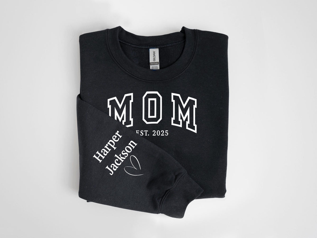 May include: Black sweatshirt with the word "MOM" in white block letters. The sleeve displays "Harper Jackson" and a heart graphic. The text "EST. 2025" is below "MOM".