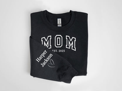 May include: Black sweatshirt with the word "MOM" in white block letters. The sleeve displays "Harper Jackson" and a heart graphic. The text "EST. 2025" is below "MOM".
