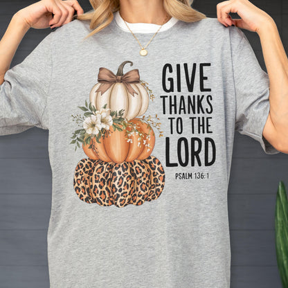 May include: A light gray t-shirt featuring a stacked pumpkin design. The pumpkins are white, orange, and leopard print, adorned with flowers and a bow. The text "GIVE THANKS TO THE LORD" is printed in black, with "PSALM 136:1" below.
