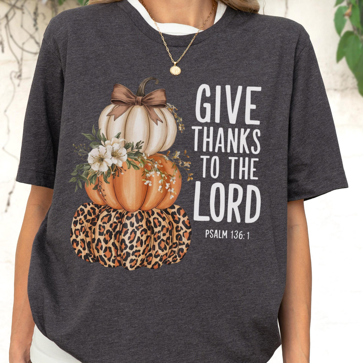 May include: Dark gray t-shirt featuring a stacked pumpkin design. The top pumpkin is white with a brown bow, the middle pumpkin is orange, and the bottom pumpkin has a leopard print. The text "GIVE THANKS TO THE LORD" is printed in white.