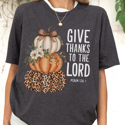 May include: Dark gray t-shirt featuring a stacked pumpkin design. The top pumpkin is white with a brown bow, the middle pumpkin is orange, and the bottom pumpkin has a leopard print. The text "GIVE THANKS TO THE LORD" is printed in white.