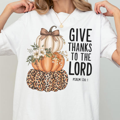 May include: White t-shirt featuring a stacked pumpkin design. The pumpkins are in shades of cream, orange, and leopard print, adorned with floral accents and a brown bow. The text "GIVE THANKS TO THE LORD" is printed in black, with "PSALM 136:1" below.