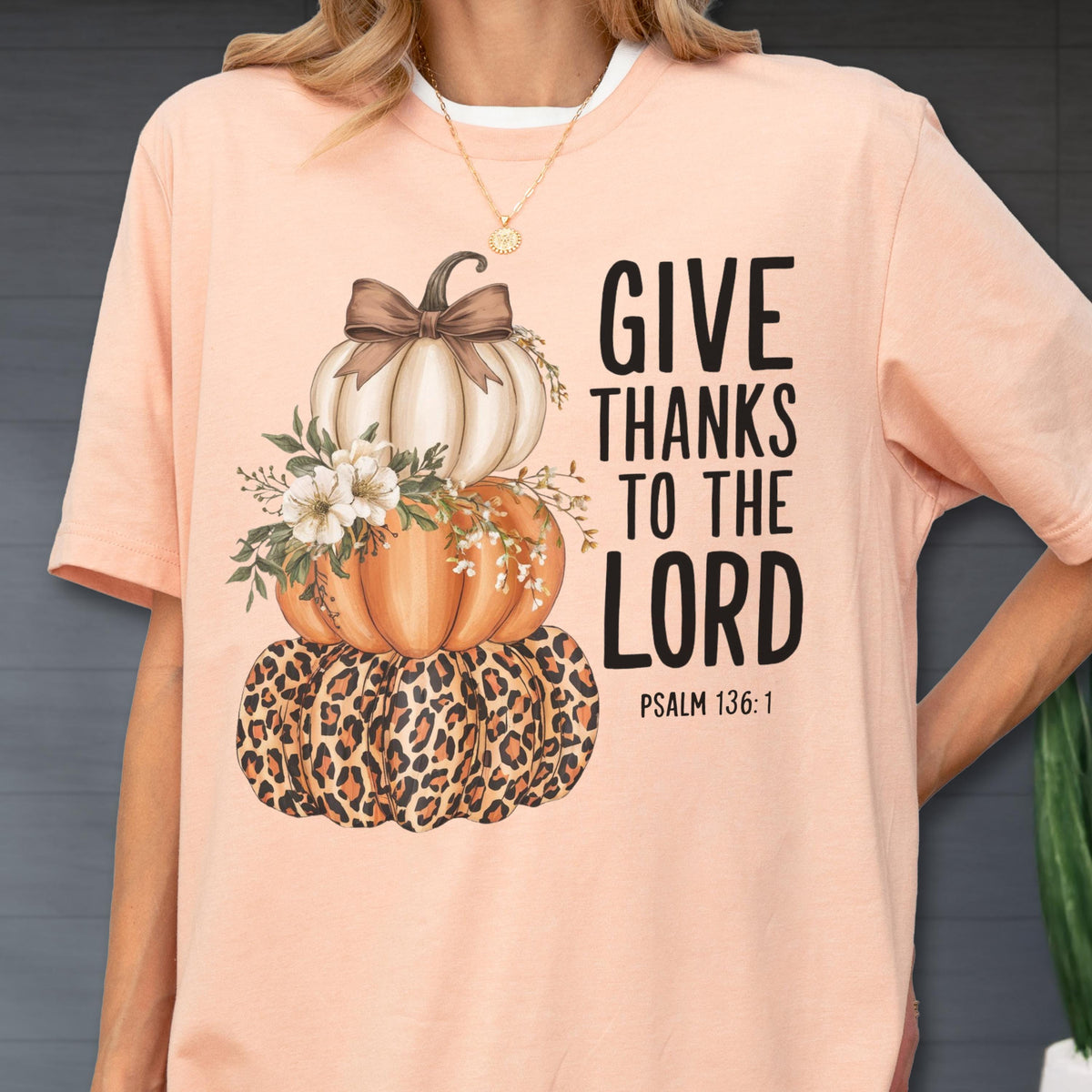 May include: Peach-colored t-shirt featuring a stacked pumpkin design with a leopard print base, orange and white pumpkins, and floral accents. The text "GIVE THANKS TO THE LORD" is printed in black, with "PSALM 136:1" below.