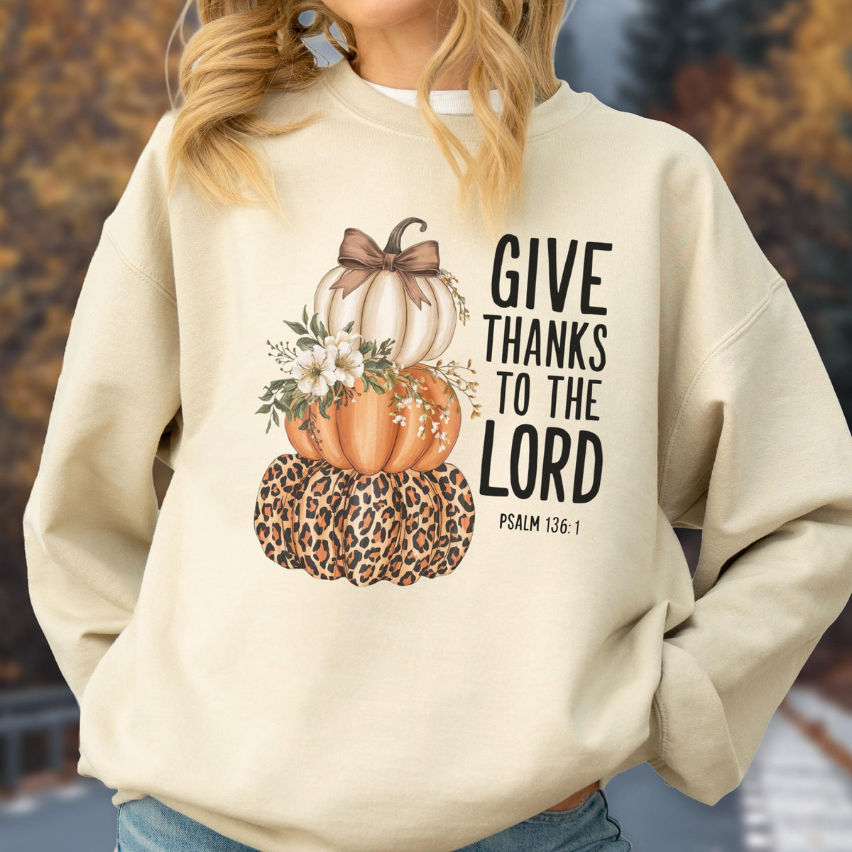 May include: Cream-colored sweatshirt featuring a stacked pumpkin design. The top pumpkin is white with a brown bow, the middle pumpkin is orange, and the bottom pumpkin has a leopard print. The text "GIVE THANKS TO THE LORD" is printed in black, with "PSALM 136:1" below.