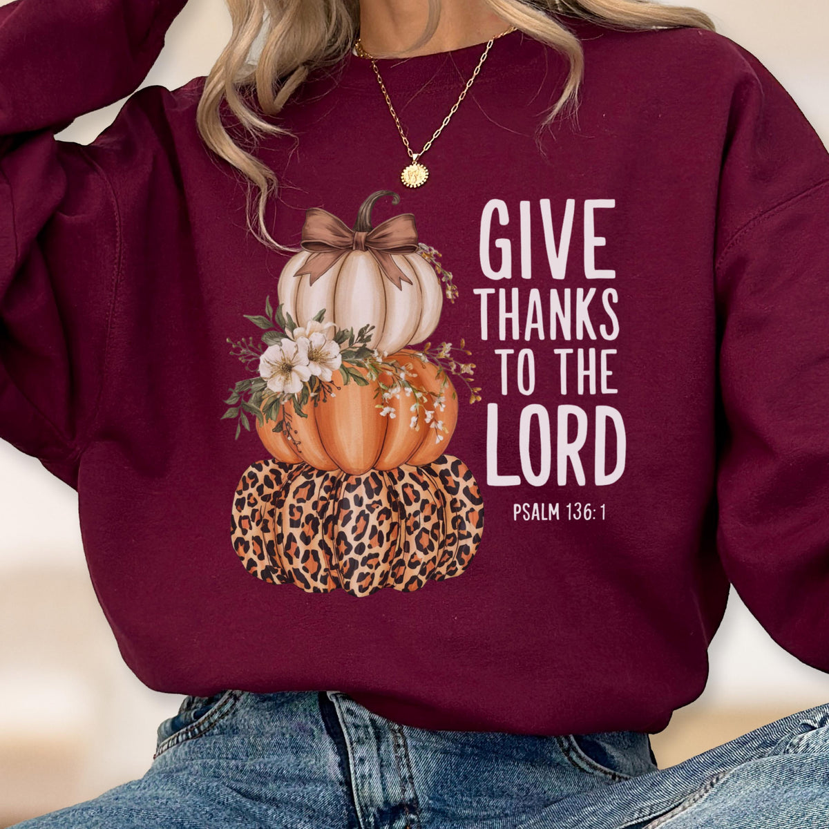 May include: Burgundy sweatshirt featuring a stacked pumpkin design. The top pumpkin is white with a brown bow, the middle is orange, and the bottom has a leopard print. Text reads "GIVE THANKS TO THE LORD" with Psalm 136:1.