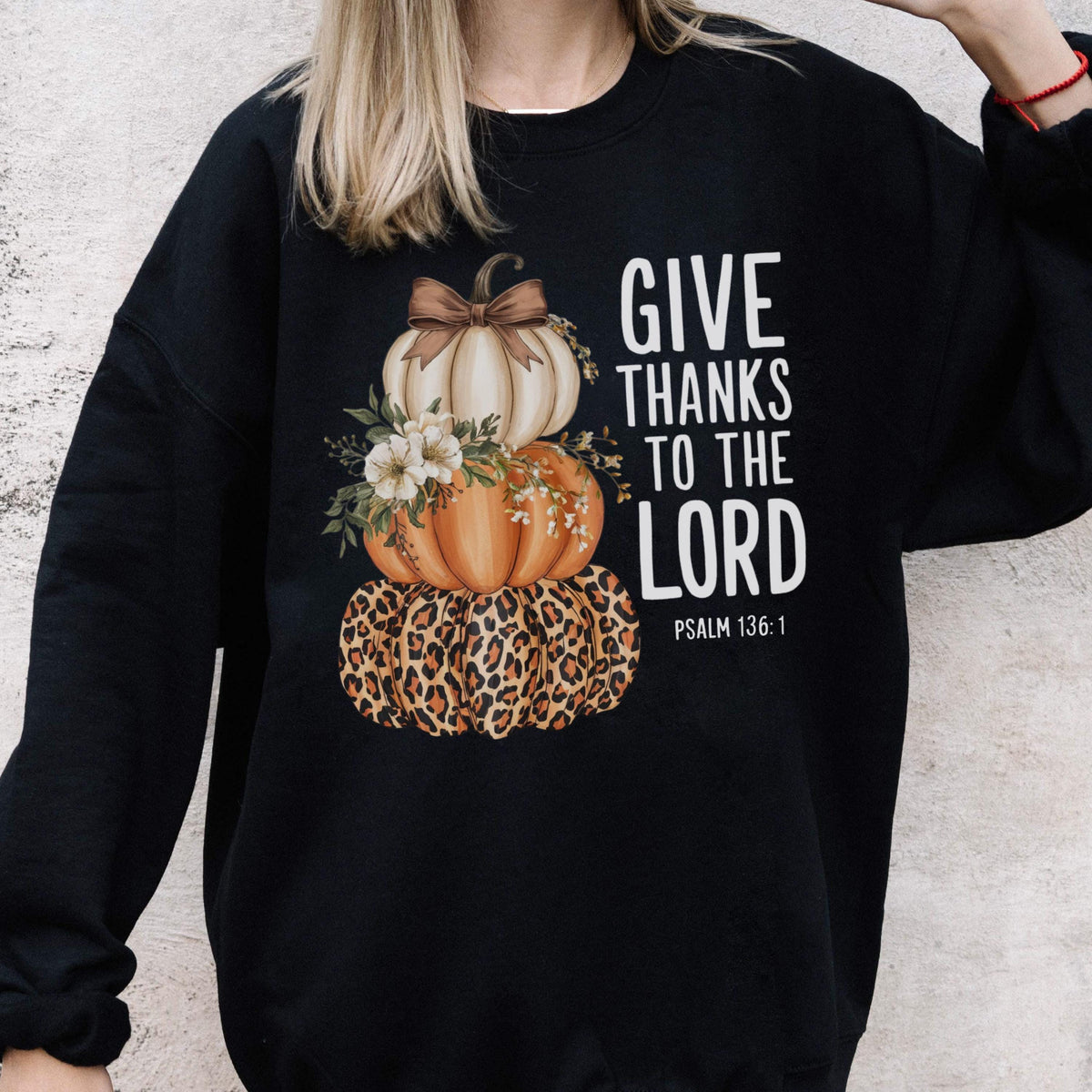 May include: Black sweatshirt featuring a stacked pumpkin design. The top pumpkin is white with a brown bow, the middle pumpkin is orange, and the bottom pumpkin has a leopard print pattern. The text "GIVE THANKS TO THE LORD" is printed in white.
