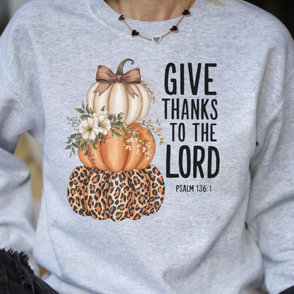 May include: A light gray sweatshirt featuring a stacked pumpkin design. The top pumpkin is white with a brown bow, the middle pumpkin is orange, and the bottom pumpkin has a leopard print. The text "GIVE THANKS TO THE LORD" is printed in black.