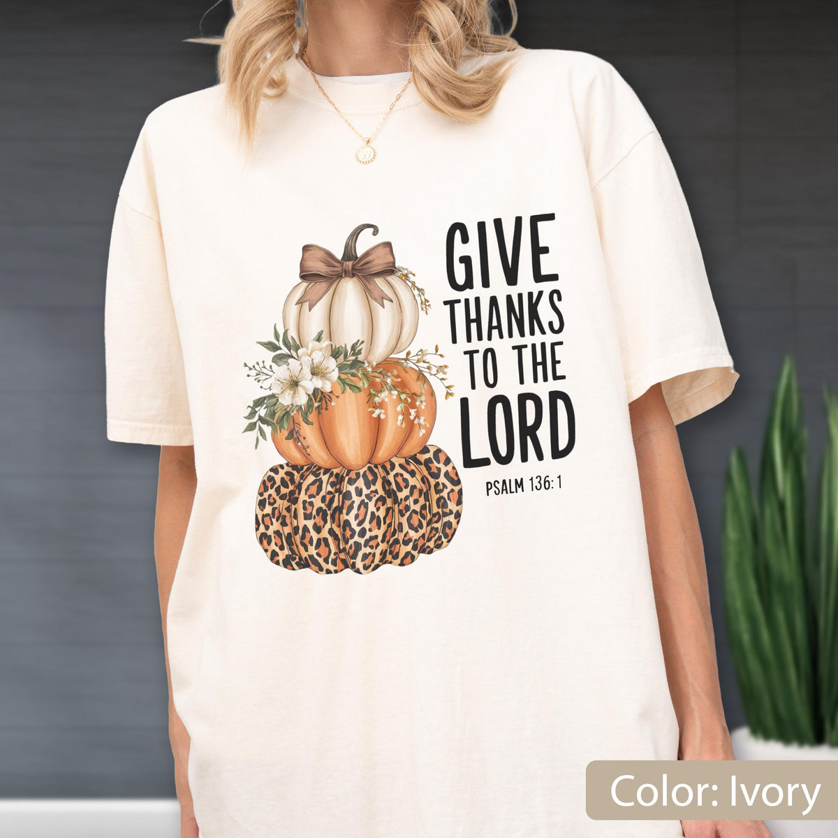 May include: Ivory-colored t-shirt featuring a stacked pumpkin design with a leopard print base, orange and white pumpkins, and floral accents. The text "GIVE THANKS TO THE LORD" is printed in black, with "PSALM 136:1" below.