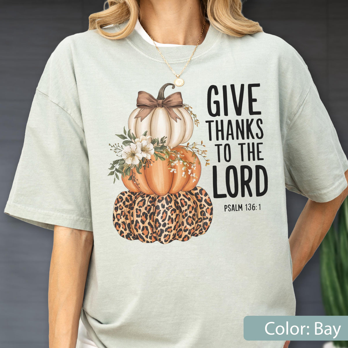 May include: A light green t-shirt featuring a stacked pumpkin design with a leopard print base, an orange middle, and a white top pumpkin. The design includes a brown bow, floral accents, and the text "GIVE THANKS TO THE LORD" with Psalm 136:1.