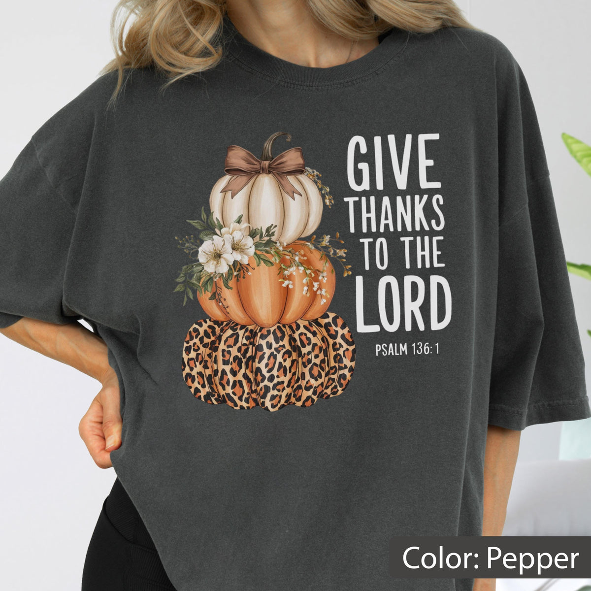 May include: Pepper-colored t-shirt featuring a stacked pumpkin design. The pumpkins are adorned with a leopard print, floral accents, and a bow. The text reads "GIVE THANKS TO THE LORD" with Psalm 136:1.