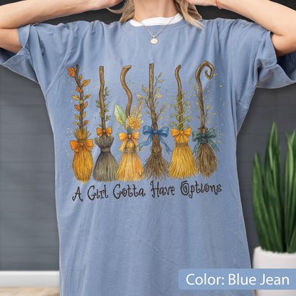 May include: A blue jean colored t-shirt featuring a graphic of six distinct broomsticks decorated with various bows, leaves, and vines. Below the brooms, the text reads A Girl Gotta Have Options in a dark, stylized, cursive-inspired font.