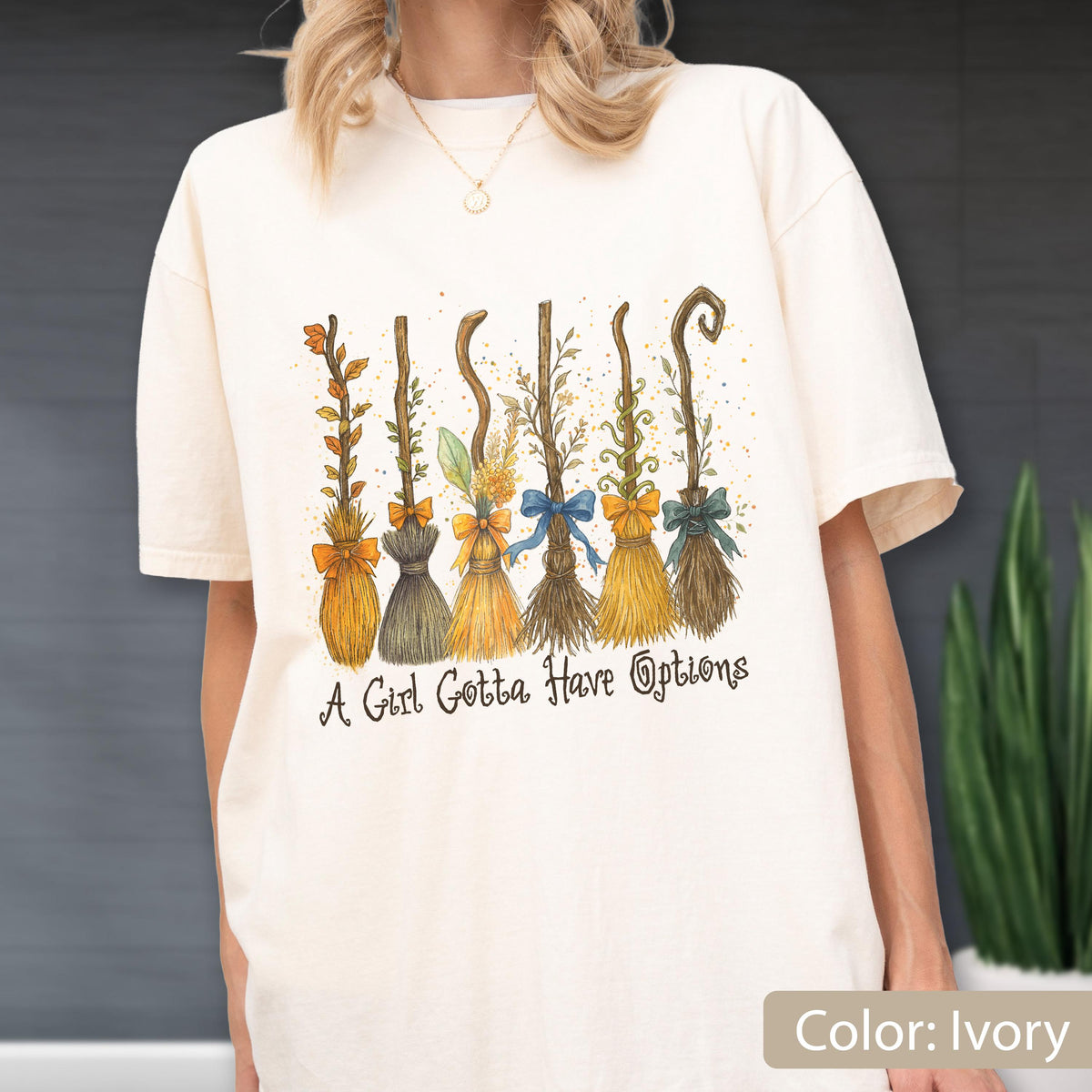 May include: An ivory t-shirt featuring a graphic print of six distinct broomsticks, each adorned with a different colored bow. Below the broomsticks, the text reads, "A Girl Gotta Have Options" in a dark, stylized, cursive-inspired font.