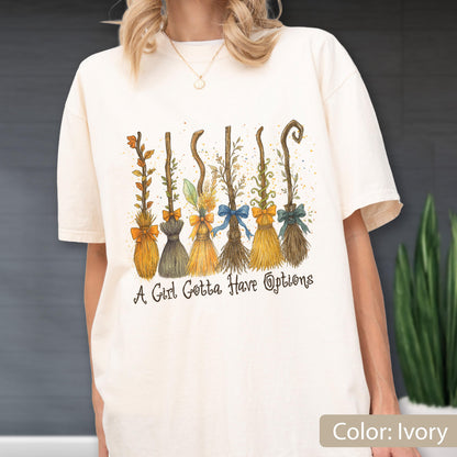May include: An ivory t-shirt featuring a graphic print of six distinct broomsticks, each adorned with a different colored bow. Below the broomsticks, the text reads, "A Girl Gotta Have Options" in a dark, stylized, cursive-inspired font.