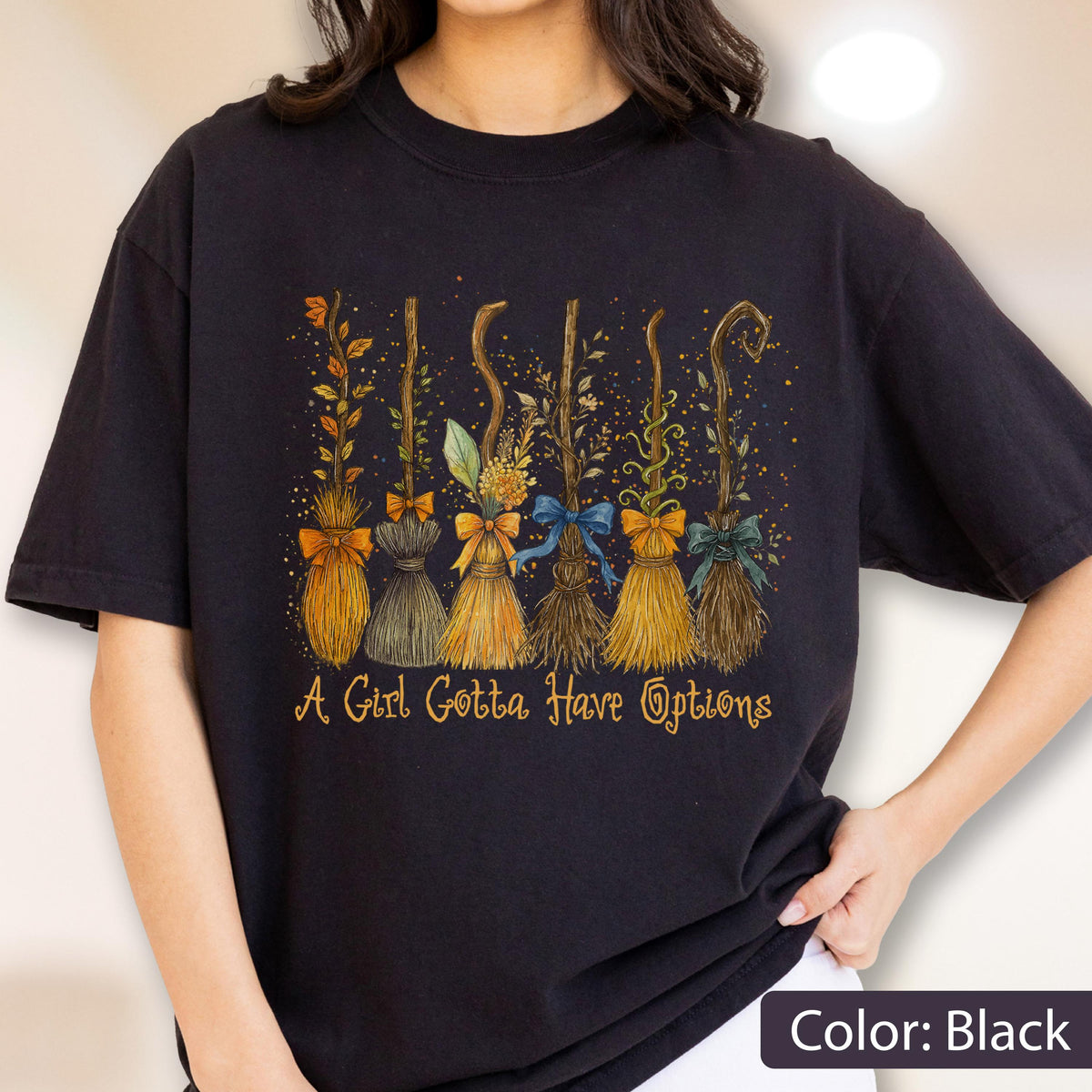 May include: A black short-sleeved t-shirt featuring a graphic of six distinct broomsticks decorated with various leaves, flowers, and ribbons. Below the brooms, the text reads A Girl Gotta Have Options in a stylized, golden-orange cursive font.