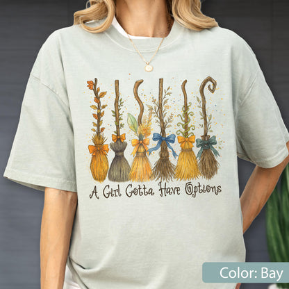May include: A light sage green t-shirt featuring a graphic of six distinct broomsticks decorated with various bows and foliage. Below the brooms, the text reads, A Girl Gotta Have Options. The shirt has a crew neckline and short sleeves.