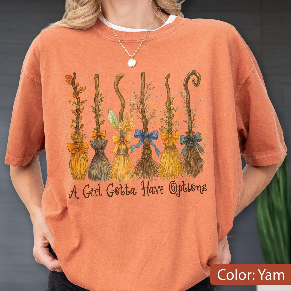 May include: A burnt orange short-sleeve t-shirt featuring a graphic of six different stylized brooms decorated with leaves, vines, and bows. Below the brooms, the text reads, A Girl Gotta Have Options, in a dark brown cursive script font.