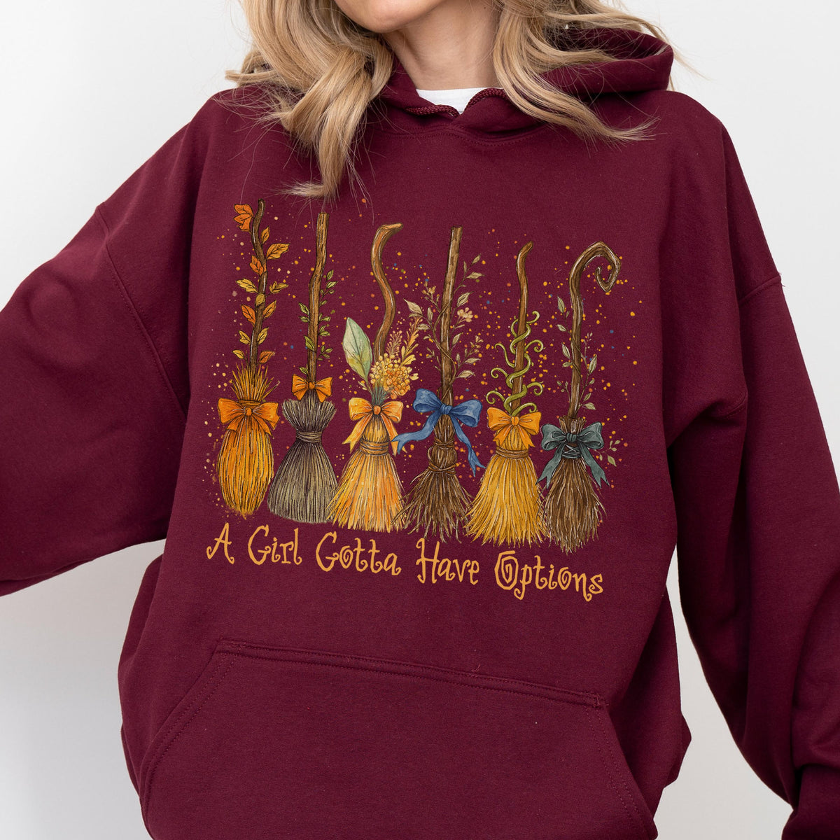 May include: A maroon hooded sweatshirt featuring a graphic print of six distinct broomsticks decorated with various bows, leaves, and vines. Below the broomsticks, the text reads, A Girl Gotta Have Options, in a stylized, cursive yellow font.