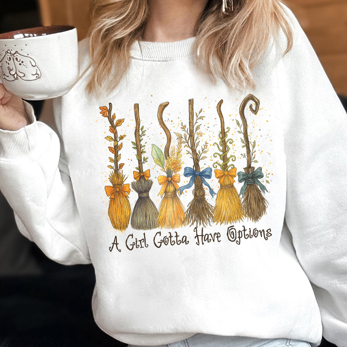 May include: A white crewneck sweatshirt featuring a graphic print of six distinct broomsticks decorated with colorful bows and foliage. Below the broomsticks, the text reads A Girl Gotta Have Options in a dark brown, stylized cursive font.