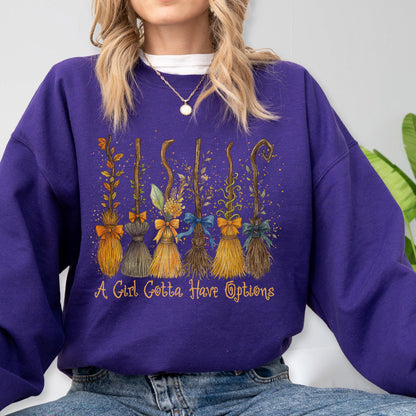 May include: A deep purple long-sleeve sweatshirt featuring a graphic print of six distinct broomsticks decorated with colorful bows and foliage. Below the brooms, the text reads A Girl Gotta Have Options in a stylized, golden-yellow cursive font.