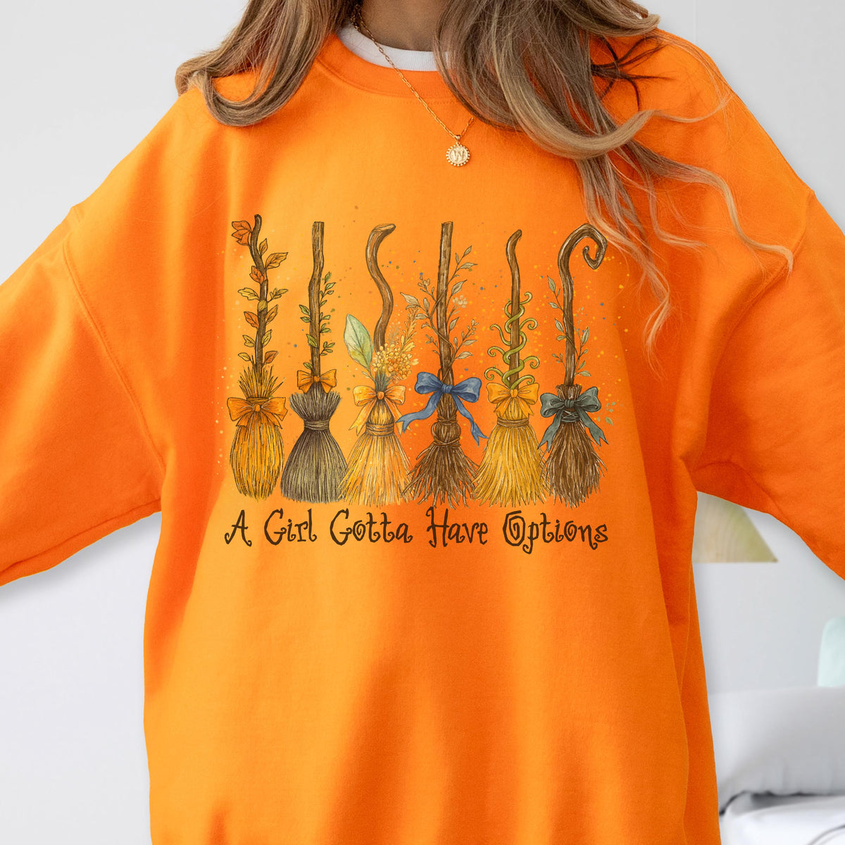 May include: A bright orange crewneck sweatshirt featuring a graphic of six distinct witch brooms adorned with colorful bows and foliage. Below the brooms, the text reads, A Girl Gotta Have Options, in a dark brown, stylized cursive font.
