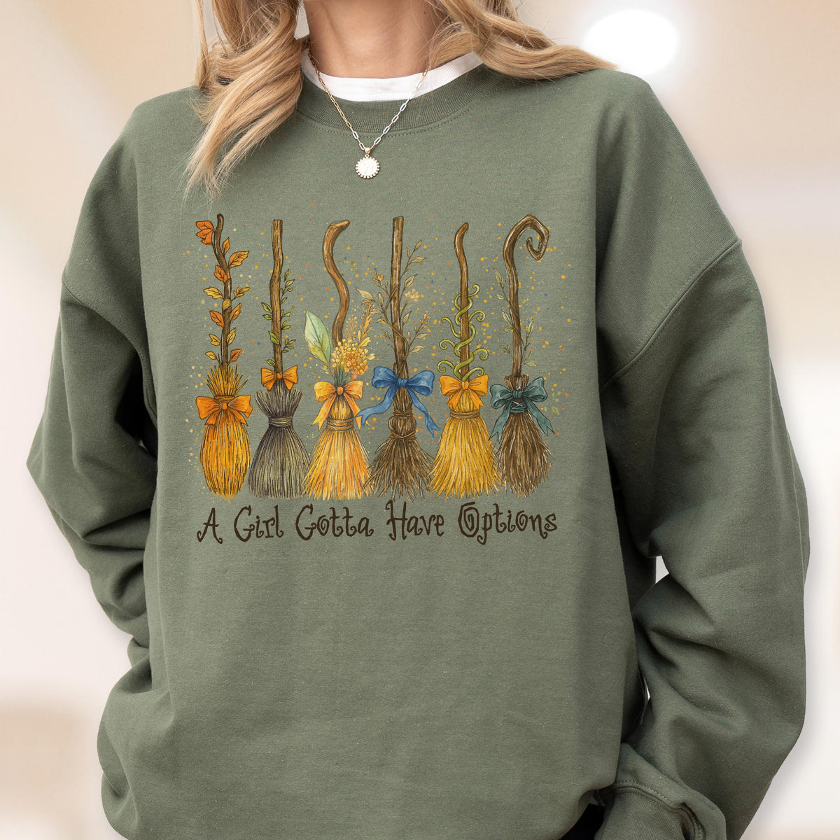 May include: An olive green crewneck sweatshirt featuring a graphic of six distinct broomsticks decorated with various bows and foliage. Below the illustration, the text reads, A Girl Gotta Have Options, in a dark brown, cursive-style font.