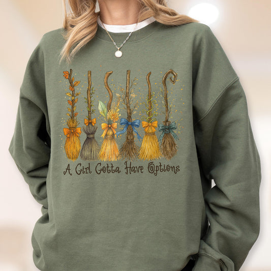 May include: An olive green crewneck sweatshirt featuring a graphic of six distinct broomsticks decorated with various bows and foliage. Below the illustration, the text reads, A Girl Gotta Have Options, in a dark brown, cursive-style font.
