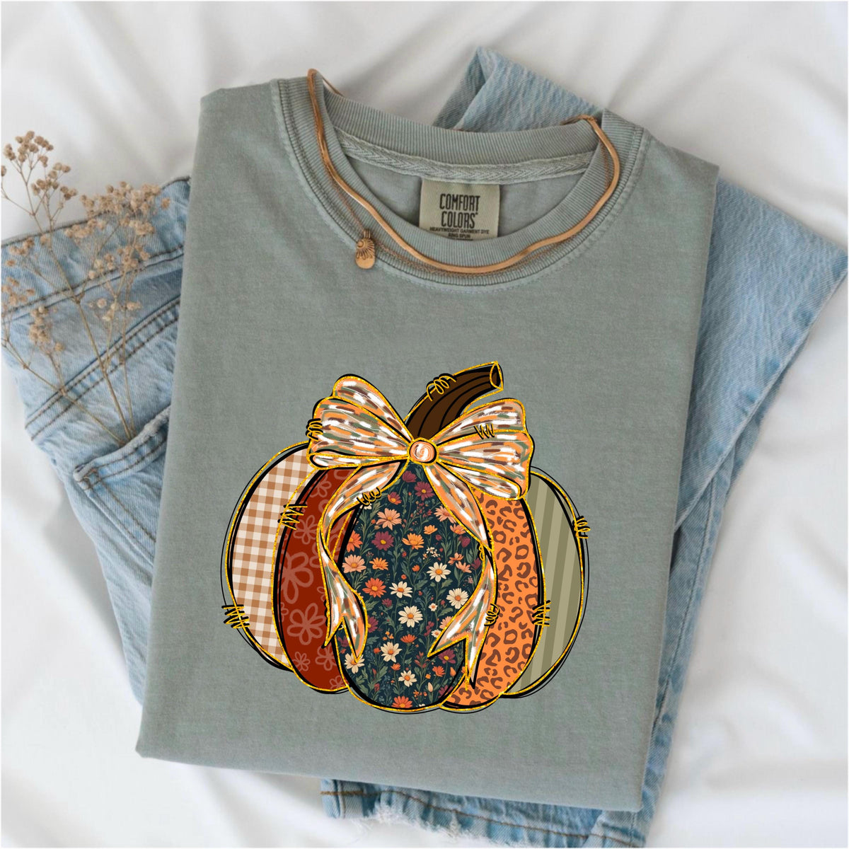 May include: Sage green Comfort Colors t-shirt with a colorful pumpkin graphic. The pumpkin design features a bow and sections with floral, plaid, and leopard print patterns. A gold necklace is draped over the shirt, which is folded on top of blue jeans.