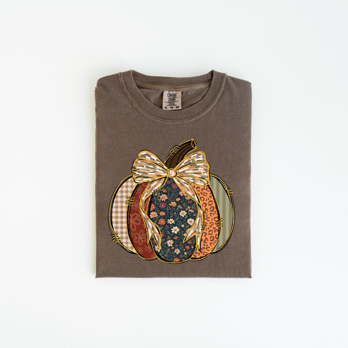 May include: A brown t-shirt with a pumpkin design. The pumpkin is composed of various patterns, including floral, plaid, and leopard print, and is topped with a decorative bow. The shirt is folded and lies flat on a white surface.