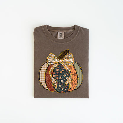 May include: A brown t-shirt with a pumpkin design. The pumpkin is composed of various patterns, including floral, plaid, and leopard print, and is topped with a decorative bow. The shirt is folded and lies flat on a white surface.