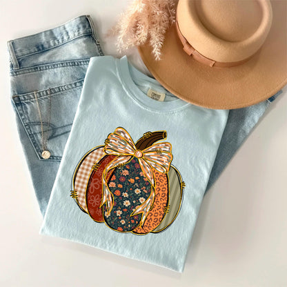 May include: A light blue t-shirt featuring a pumpkin design with a bow. The pumpkin is composed of various patterns, including floral, plaid, and leopard print. A pair of blue jeans and a tan hat are also in the image.