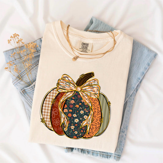 May include: Cream-colored Comfort Colors t-shirt featuring a pumpkin design. The pumpkin is segmented with various patterns, including floral, plaid, and leopard print, and is topped with a large bow. A gold necklace rests on the shirt.