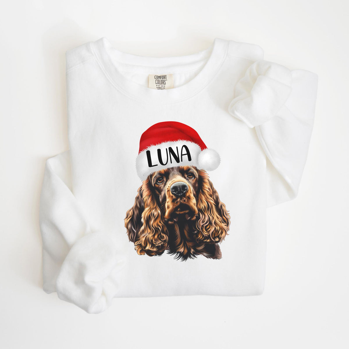 May include: White sweatshirt featuring a detailed illustration of a Cocker Spaniel wearing a Santa hat. The hat is red with a white trim and the name "LUNA" in black letters. The dog has brown fur.