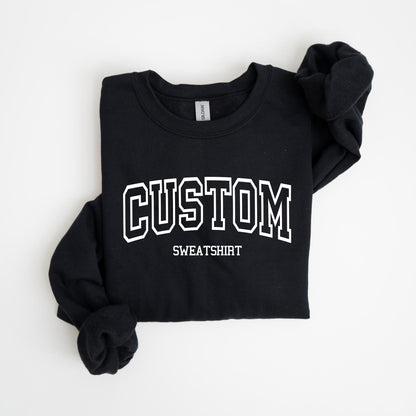 May include: Black sweatshirt with the word "CUSTOM" in white, outlined block letters. Below the word "CUSTOM" is the word "SWEATSHIRT" in a smaller font. The sweatshirt is folded neatly on a white surface.