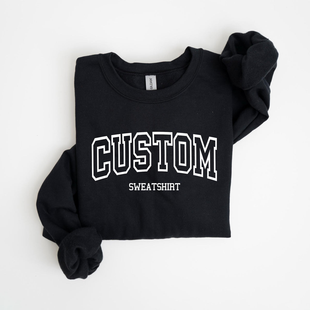 May include: Black sweatshirt with the word "CUSTOM" in white, collegiate-style lettering. Below the word "CUSTOM" is the word "SWEATSHIRT" in a smaller font. The sweatshirt is folded neatly on a white surface.