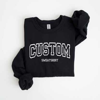 May include: Black sweatshirt with the word "CUSTOM" in white, collegiate-style lettering. Below the word "CUSTOM" is the word "SWEATSHIRT" in a smaller font. The sweatshirt is folded neatly on a white surface.