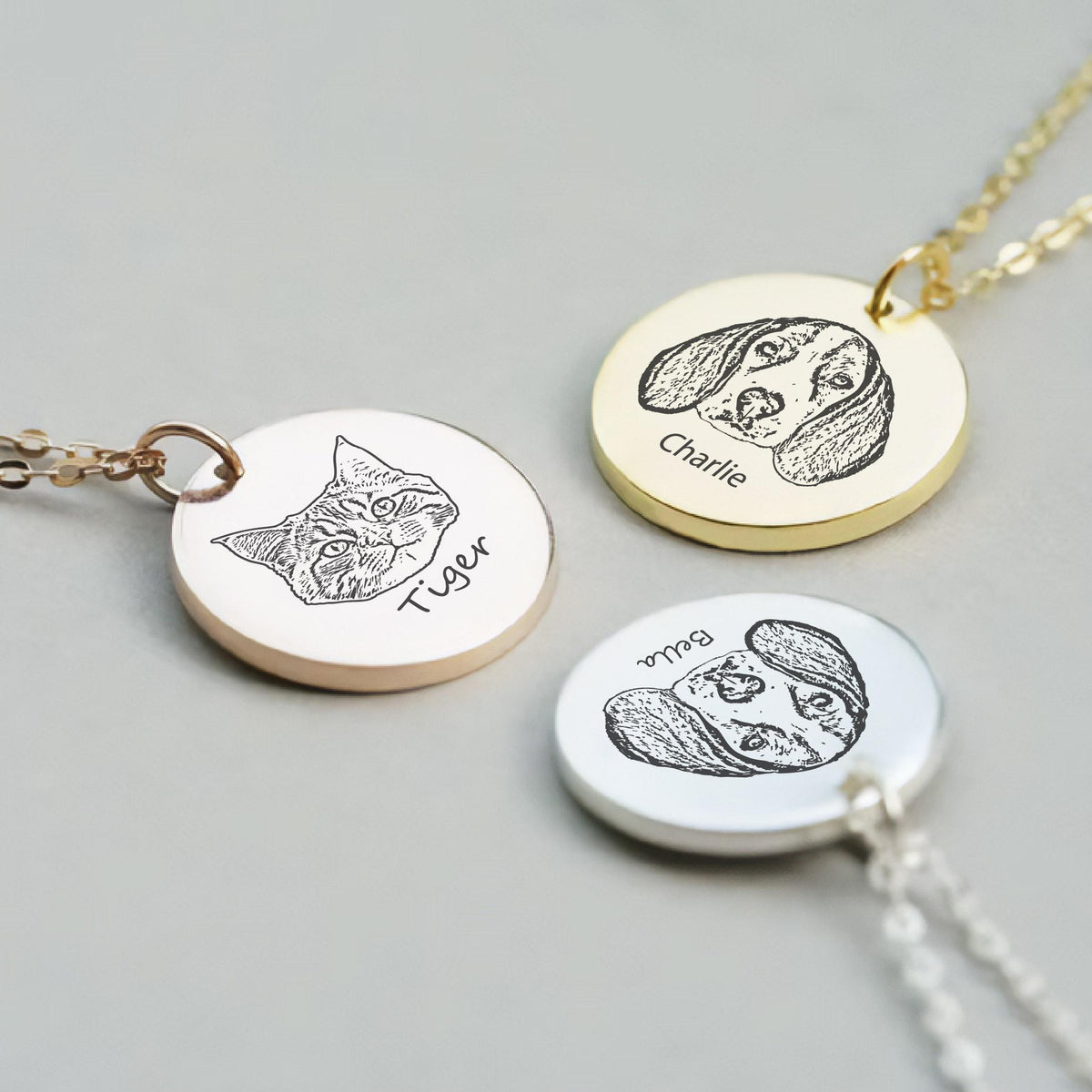 May include: Three personalized pet portrait necklaces. One rose gold necklace with a cat portrait and the name "Tiger". A gold necklace with a dog portrait and the name "Charlie". A silver necklace with a dog portrait and the name "Bella".