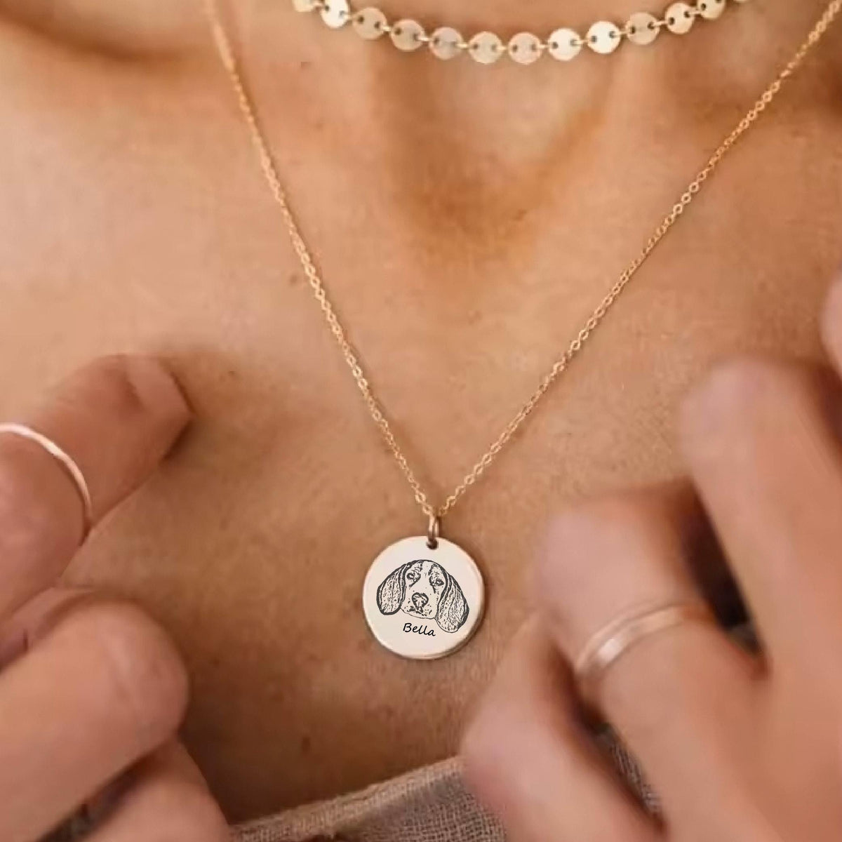 May include: Gold-colored necklace with a round pendant featuring a sketched dog portrait and the name "Bella". The necklace has a delicate chain and is worn around the neck. A matching gold choker is also visible.