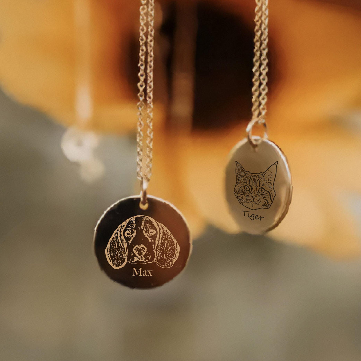 May include: Two gold-colored pendant necklaces. One pendant is round with a dog portrait and the name "Max" engraved. The other pendant is oval with a cat portrait and the name "Tiger" engraved. Both pendants hang from delicate gold chains.