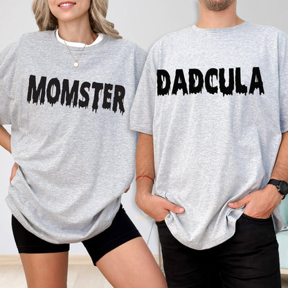 May include: Two light gray crew neck t-shirts. The left shirt features the black text MOMSTER in a dripping font. The right shirt features the black text DADCULA in a dripping font. Both shirts have short sleeves and a standard fit.