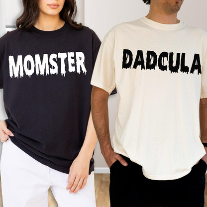 May include: A black t-shirt with the white text MOMSTER in a dripping font and a cream-colored t-shirt with the black text DADCULA in a dripping font. Both shirts feature short sleeves and a crew neckline, displaying a graphic design on the chest.