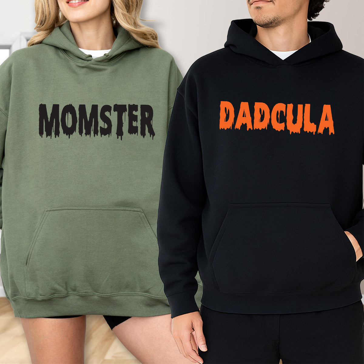 May include: Two hooded sweatshirts, one olive green and one black. The olive green sweatshirt has the word "MOMSTER" printed in black, and the black sweatshirt has the word "DADCULA" printed in orange. Both sweatshirts have a front pocket.