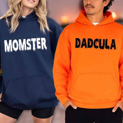 May include: Two hooded sweatshirts, one navy blue and one orange. The navy blue sweatshirt has the word "MOMSTER" in white, dripping font. The orange sweatshirt has the word "DADcula" in black, dripping font. Both have a front pocket.