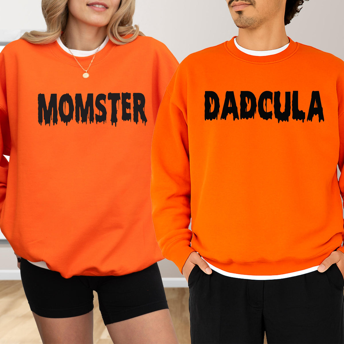 May include: Two orange sweatshirts with black text. One says "MOMSTER" and the other says "DADCULA". Both sweatshirts have a white crew neck collar. The sweatshirts are worn with black shorts and pants.